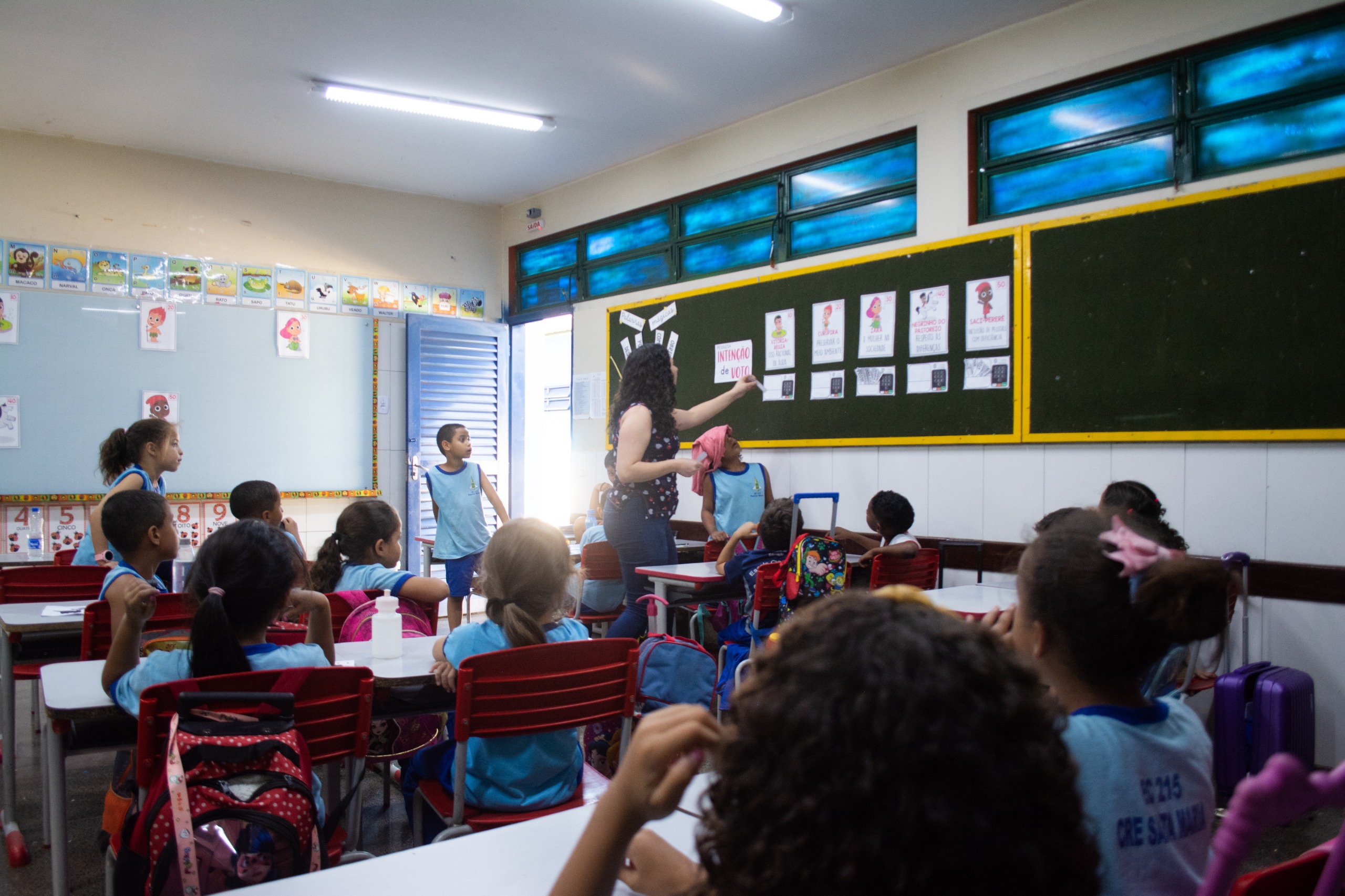 Imagem de uma sala de aula com a professora na frente de um quadro verde